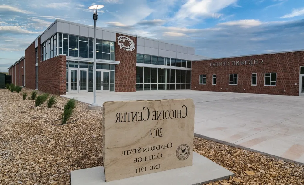 An athletic building of brick, metal, and glass, with a sign in the foreground reading Chicoine Center, 2014, Chadron State College, Est. 1911